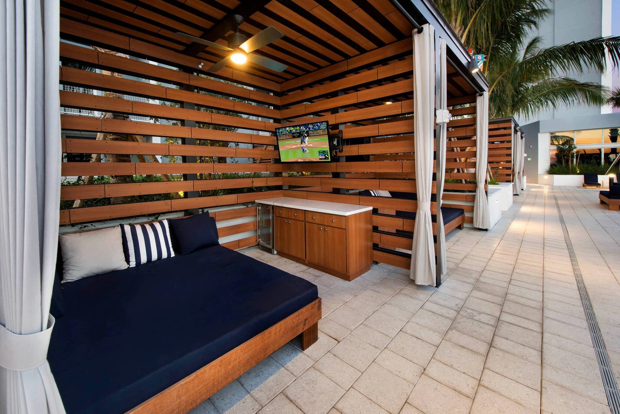Rooftop lounge with horizontal wood slat walls, built-in navy seating, wooden bar counter, wall-mounted TV, and palm trees visible in background.