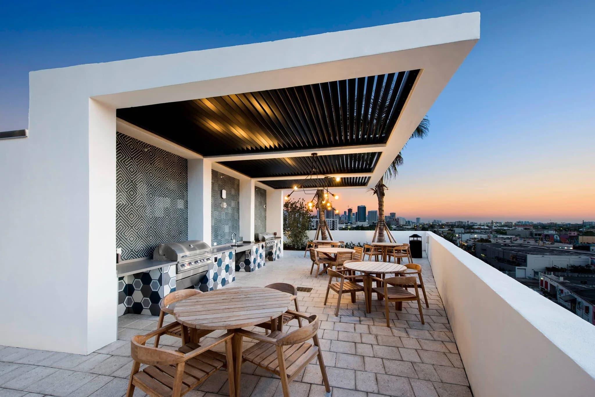 Rooftop terrace with wooden dining tables, built-in grill, louvered pergola, and Miami skyline view at dusk.