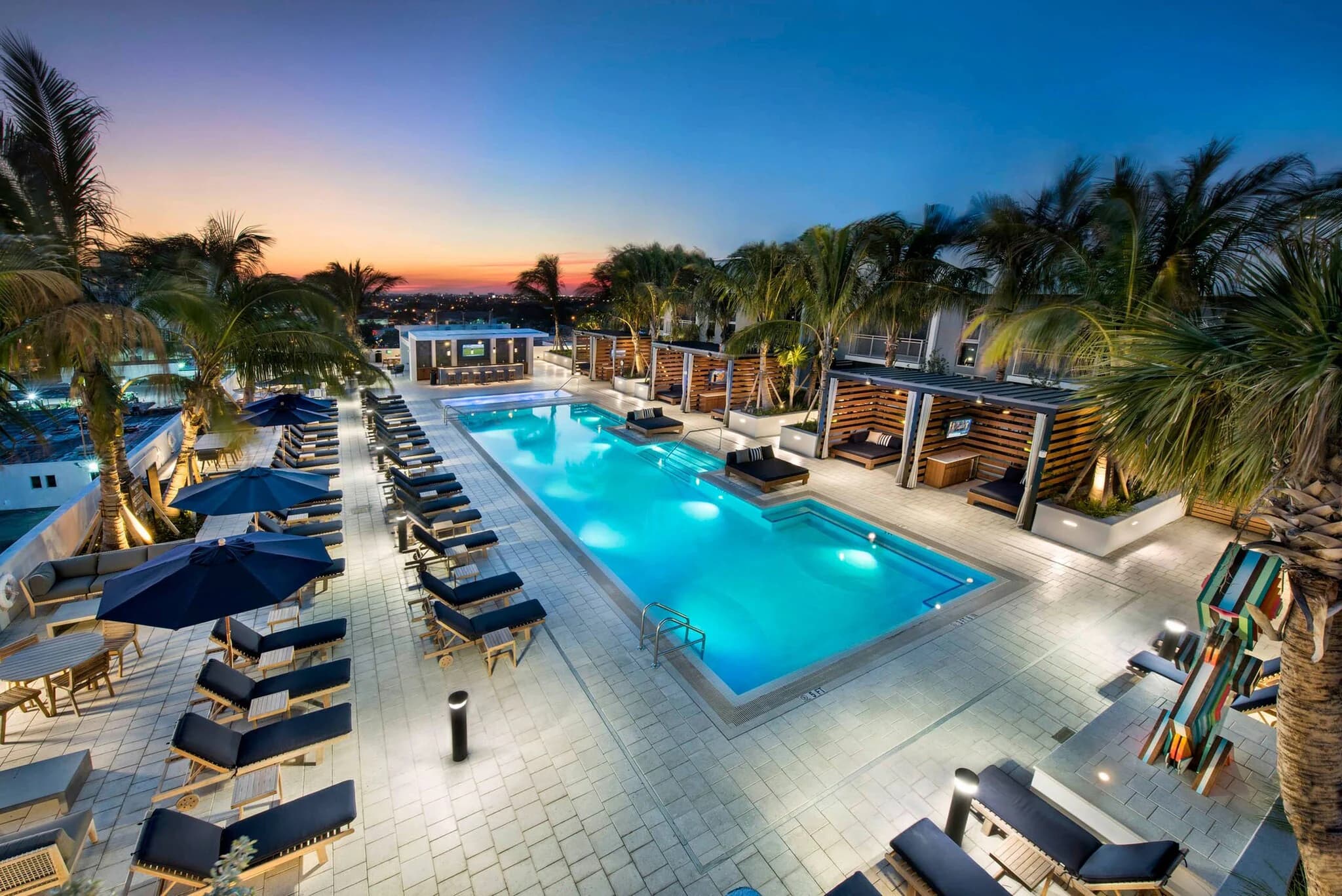 Resort pool at dusk surrounded by lounge chairs, palm trees, wooden cabanas, and illuminated patio at AMLI Midtown 29.