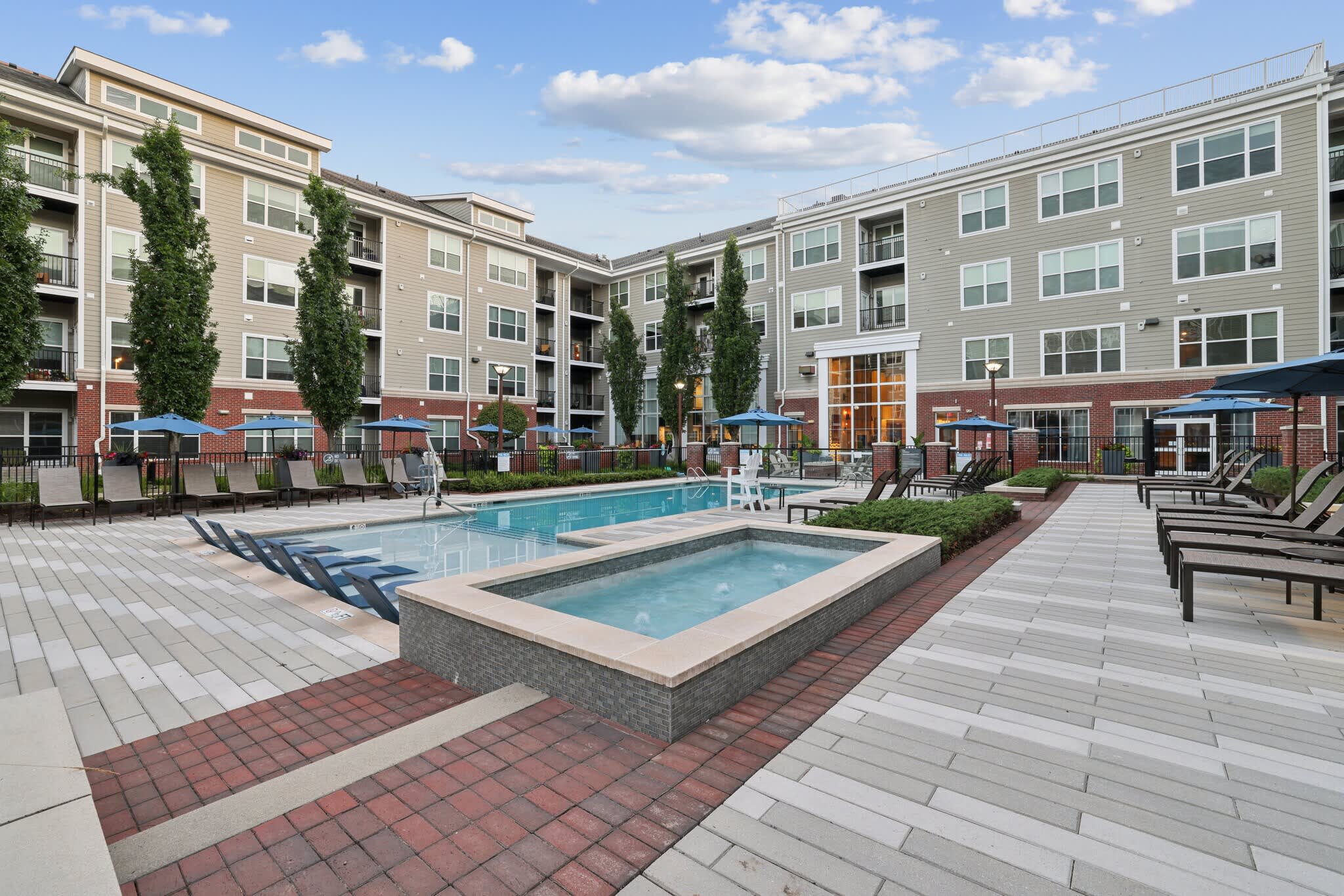 Resort-style courtyard with two pools, hot tub, lounge chairs, and blue umbrellas, flanked by multi-story apartment buildings with brick and siding exterior.