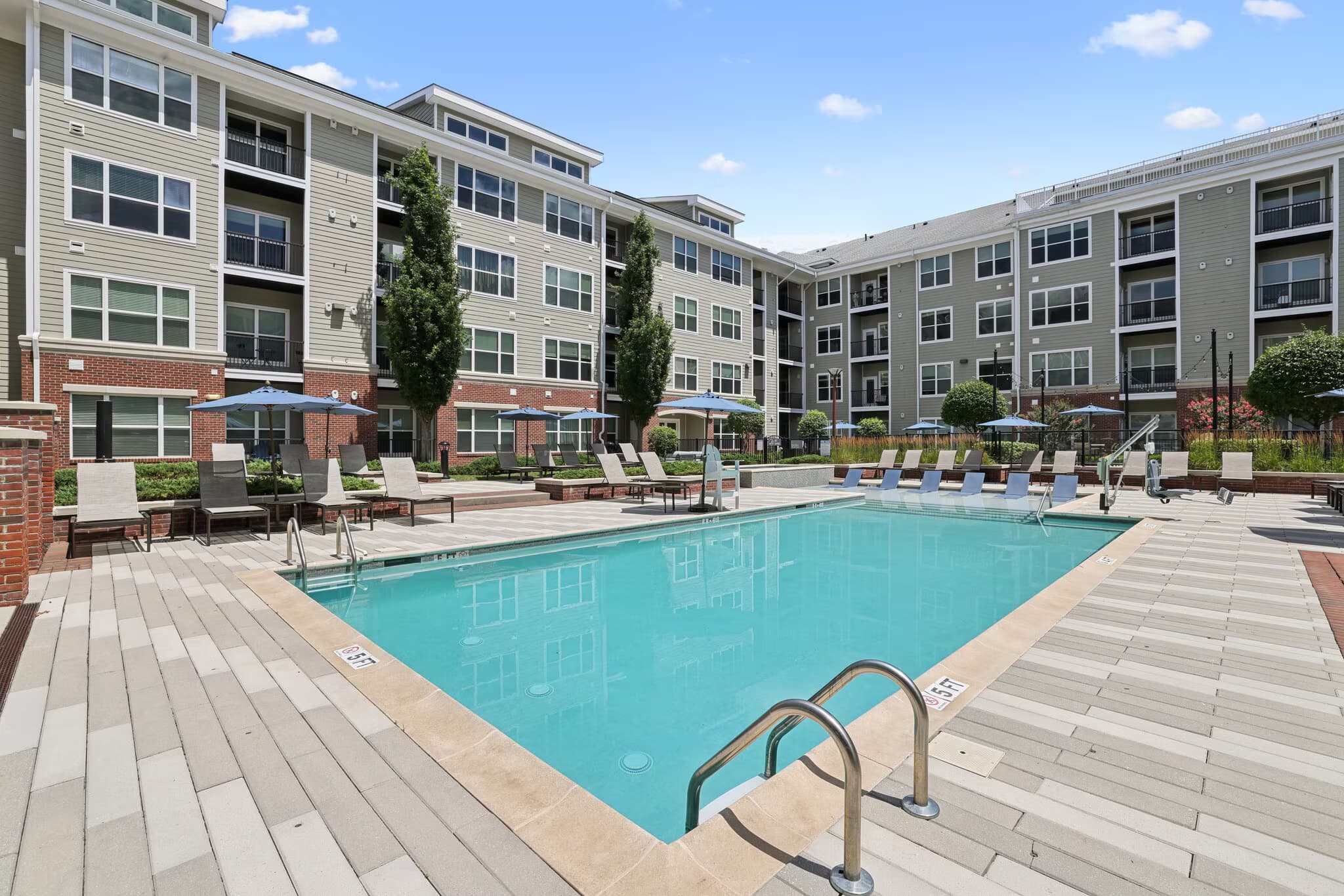 Outdoor swimming pool with lounge chairs and umbrellas, surrounded by multi-story residential buildings with brick and siding facades.