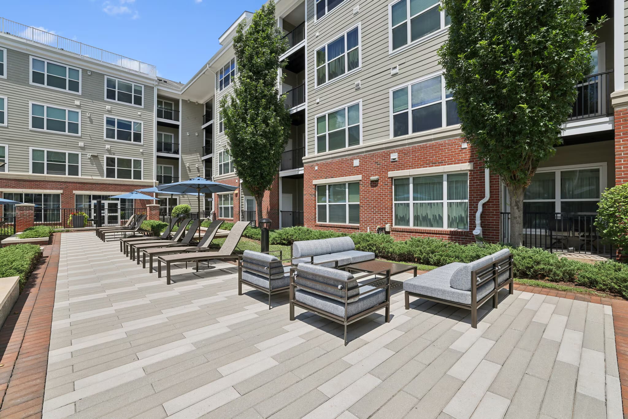 Outdoor courtyard with gray patio seating, upholstered lounge chairs, and ivy-covered brick apartment buildings under clear sky.