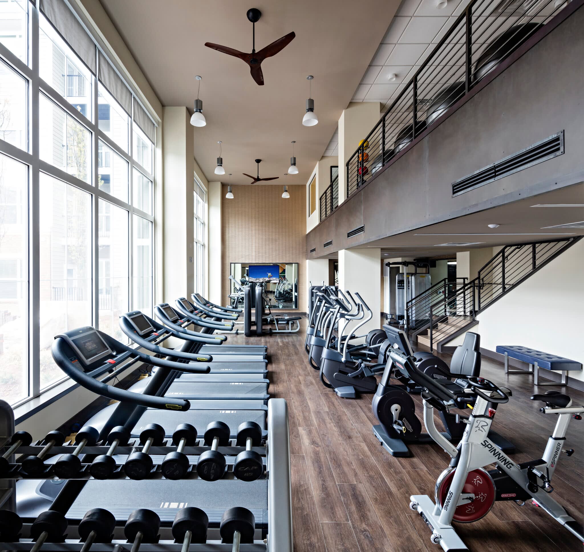 Modern gym with rows of treadmills and exercise bikes, dumbbell rack, two-story space with large windows, pendant lights, and ceiling fans.