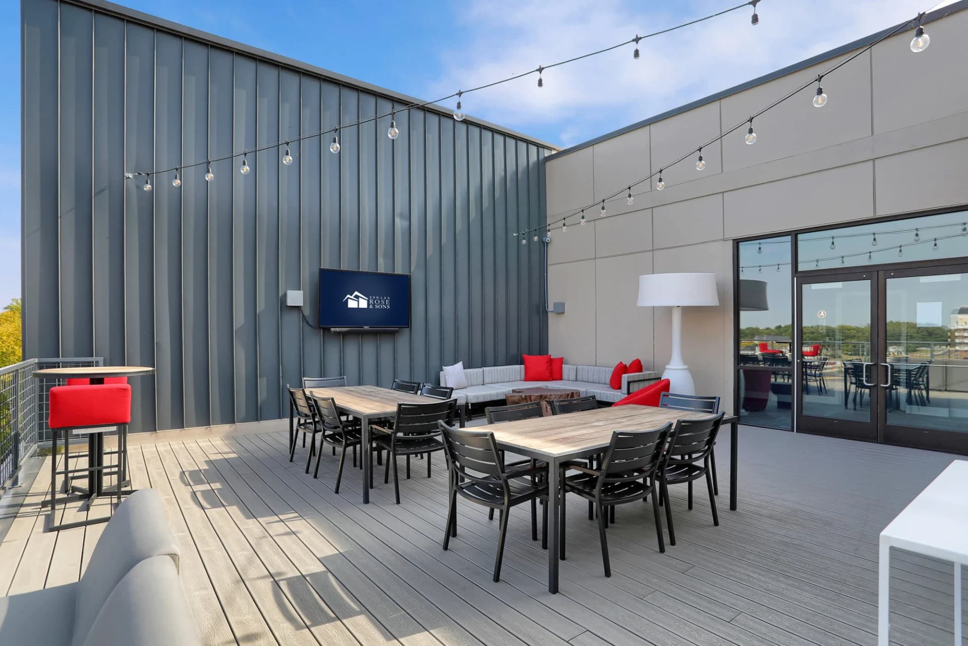 Rooftop terrace with composite decking, dining tables, gray sofa with red pillows, pendant lighting, and glass doors to interior space.
