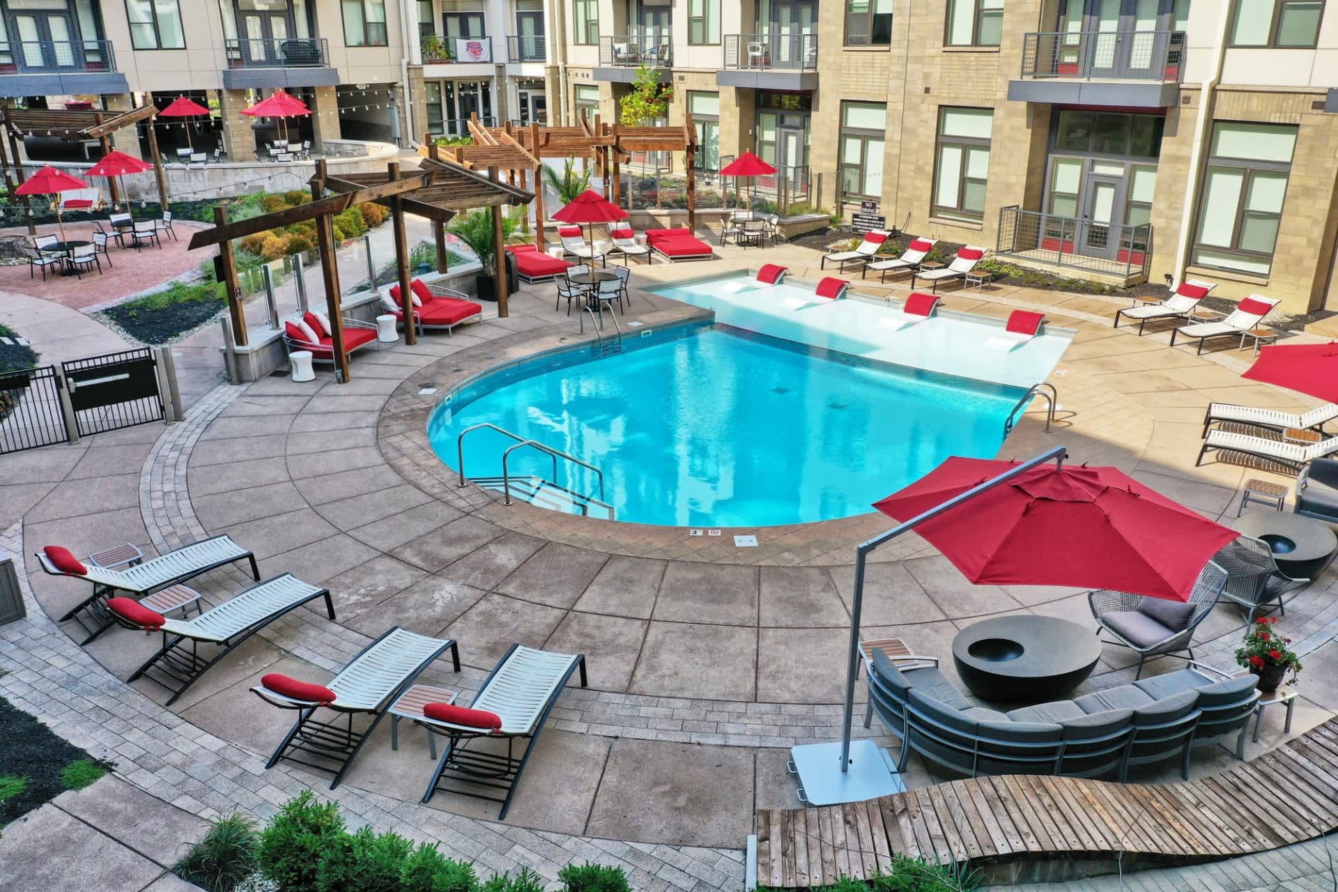 Aerial view of curved swimming pool surrounded by red umbrellas, lounge chairs, wooden pergolas, and fire pit in courtyard of multi-story residential building.