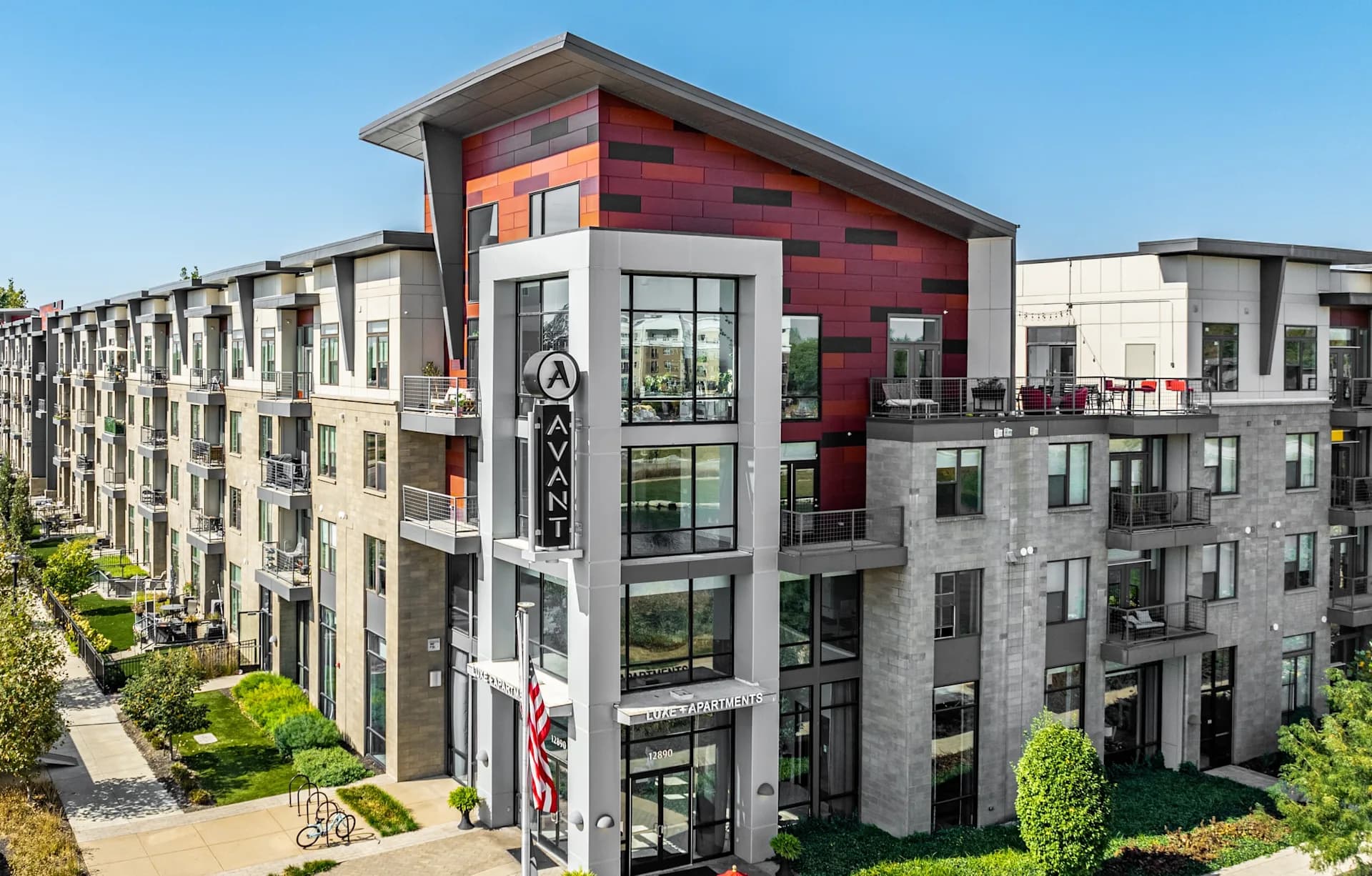 Modern multi-story apartment building with red brick and white facade, ground-floor retail, landscaped courtyard with green shrubs.