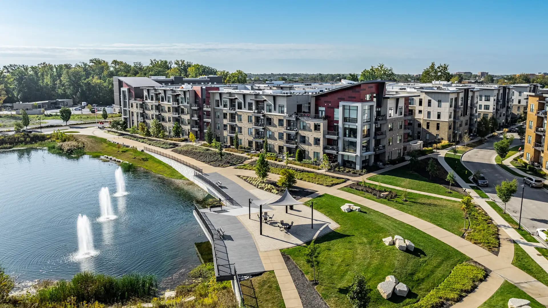 Waterfront apartment complex with multi-story buildings in varied colors, paved pathways, landscaped lawn, and pond with fountains.