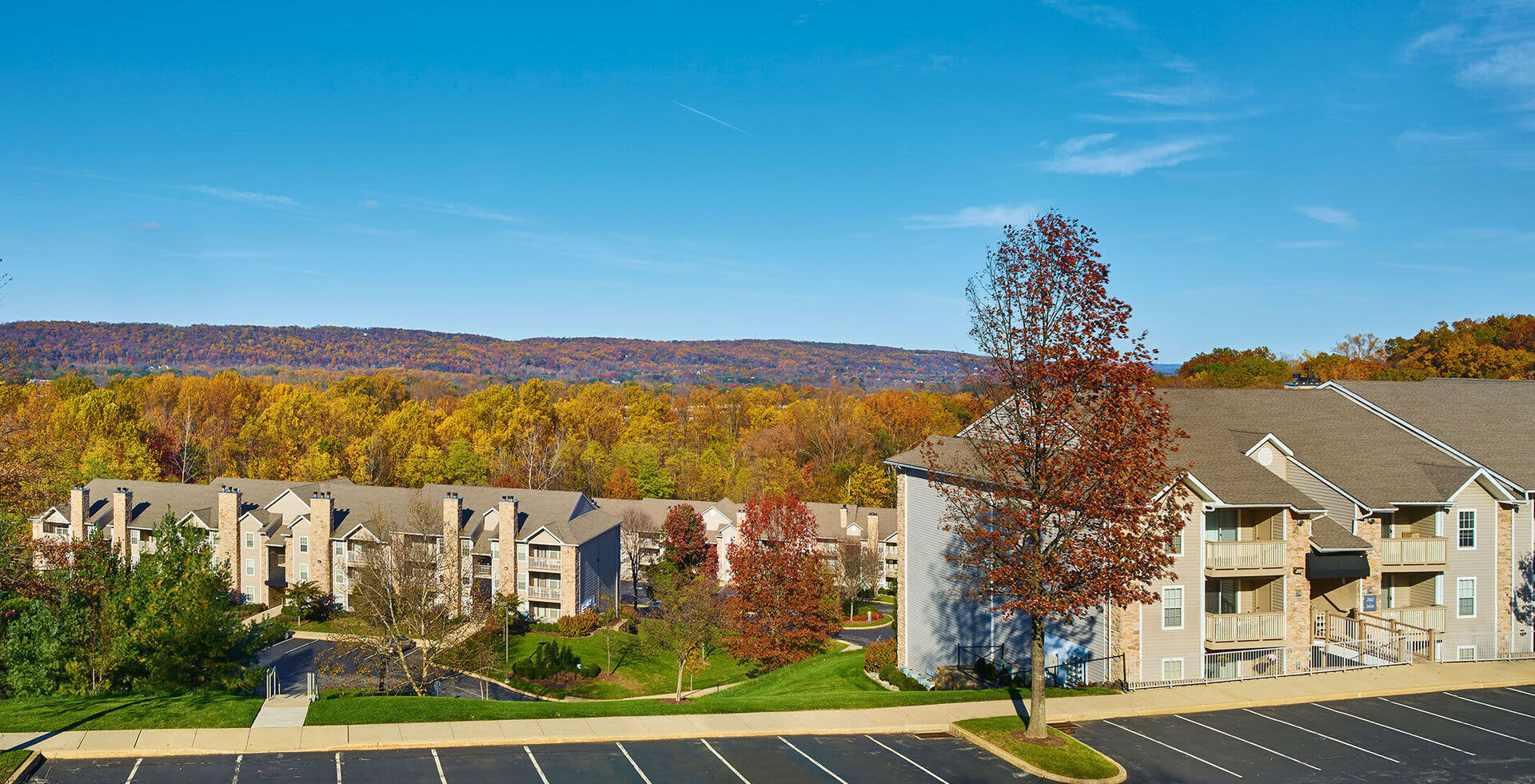 Multi-story apartment complex with tan and white buildings, tree-lined grounds with fall foliage, and forested hills in background.