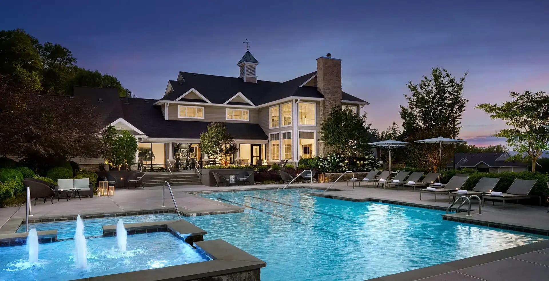 Two-story colonial home with black roof and brick chimney overlooking lit swimming pool and hot tub with lounge chairs at twilight.