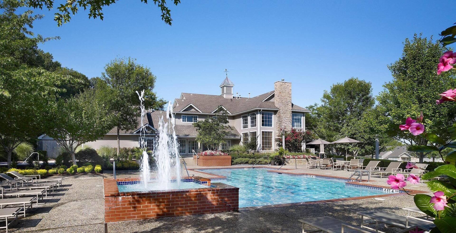 Resort-style swimming pool with brick fountain, lounge chairs, and two-story residential building with stone and brick exterior in background.