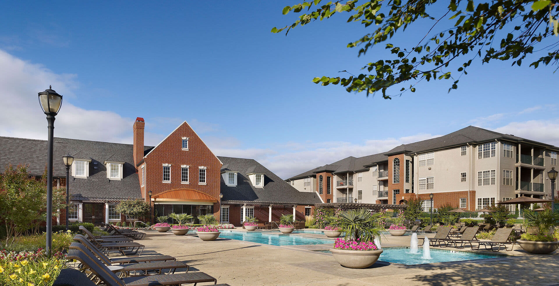 Resort-style pool courtyard with lounge chairs, pink flowering planters, brick residential buildings, and clear blue sky.