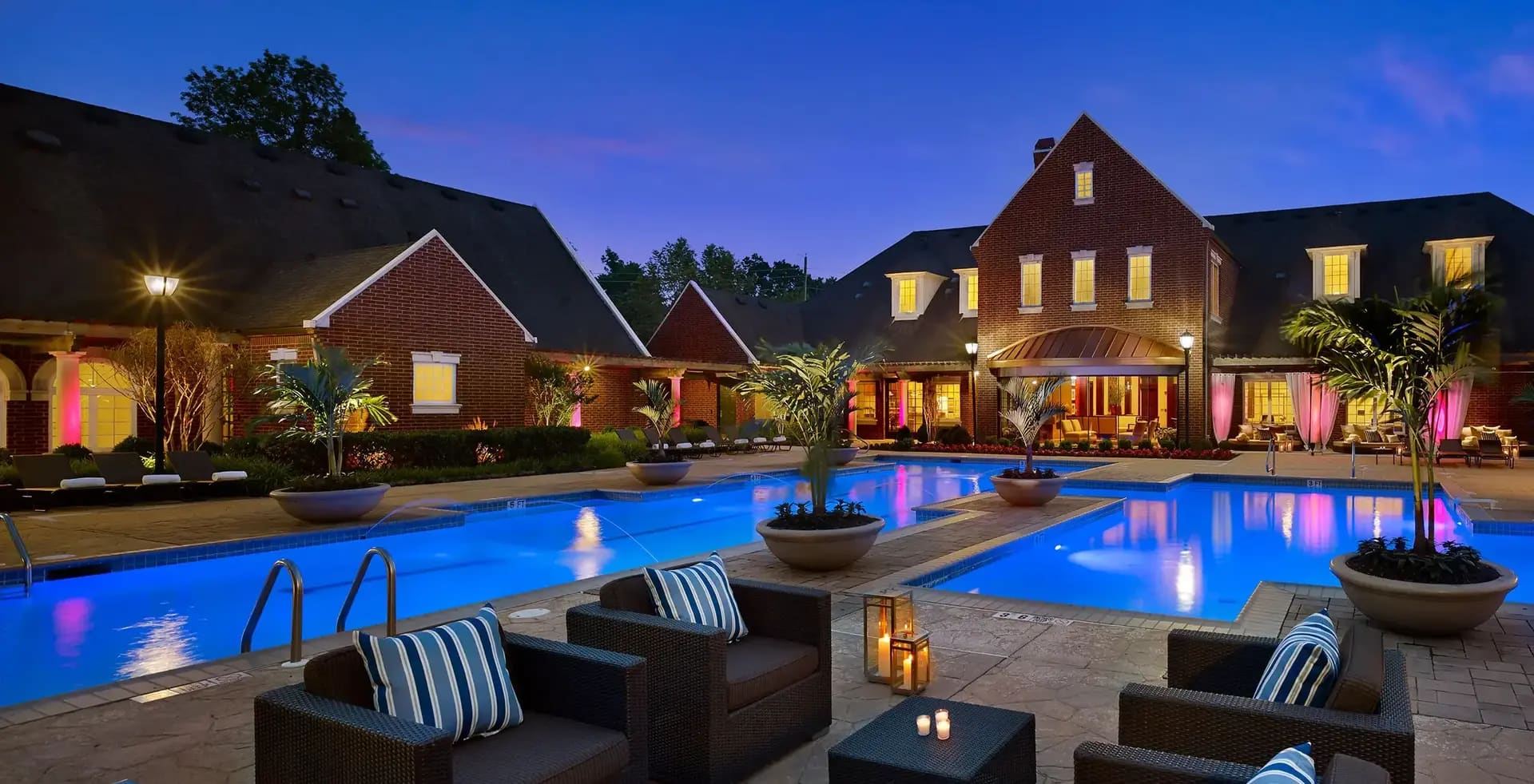 Nighttime view of large lit swimming pool with lounge chairs, fire bowls, and brick residential buildings with lit windows in background.