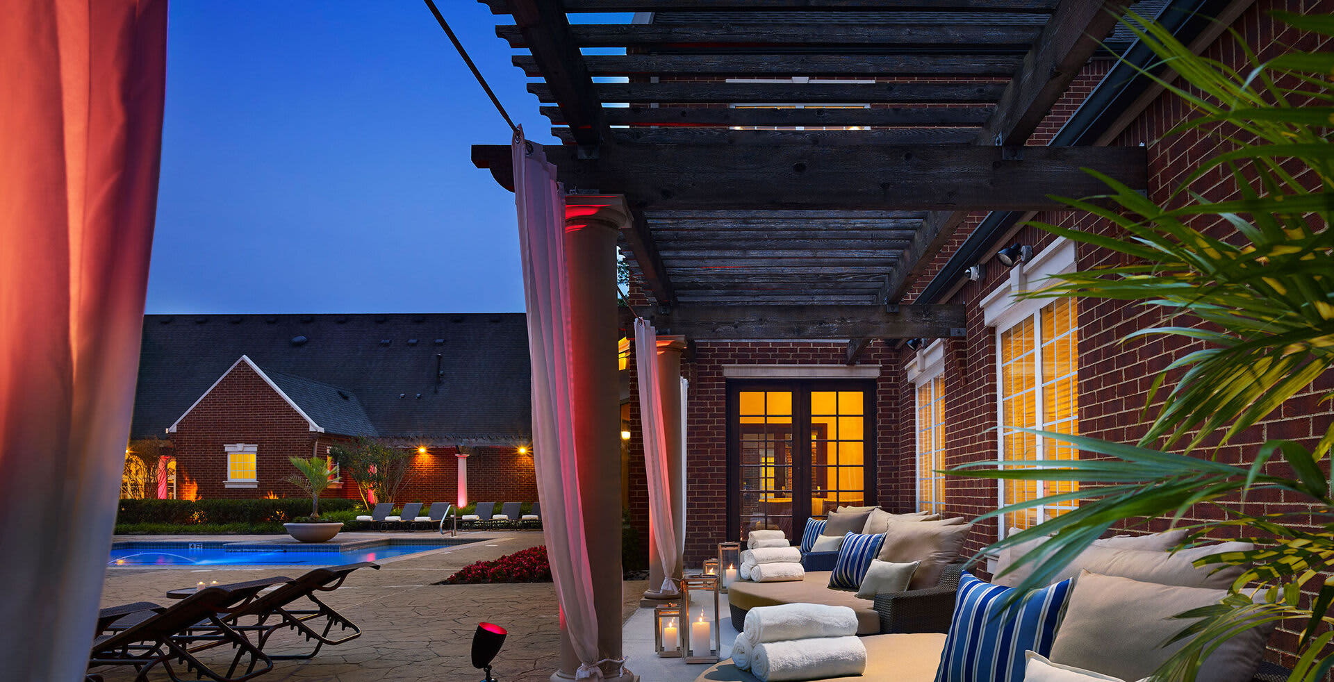 Covered patio with lounge seating, striped pillows, potted plants, and pergola overlooking lit pool and brick building at twilight.