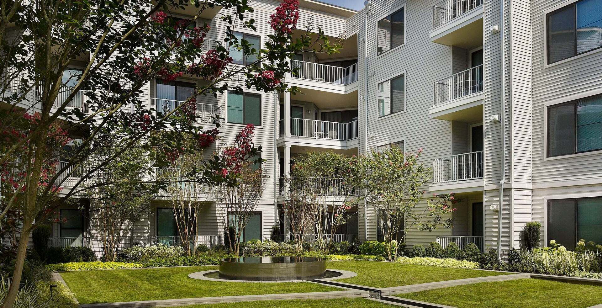 Courtyard with circular water feature, green lawn, flowering trees with red blooms, and multi-story white residential buildings with balconies.