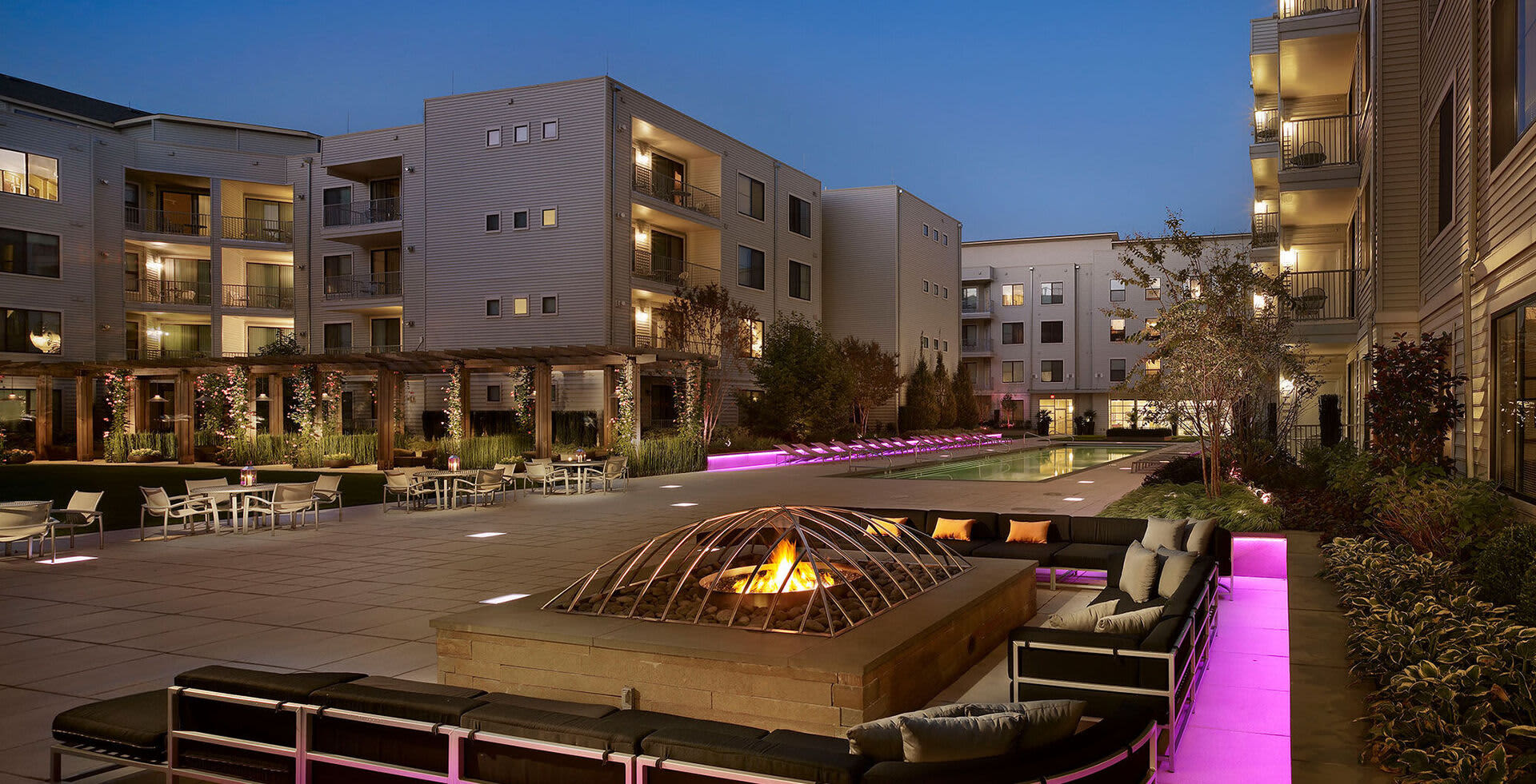 Courtyard at dusk with lit fire pit, modern seating areas, pink accent lighting, and multi-story residential buildings surrounding the space.