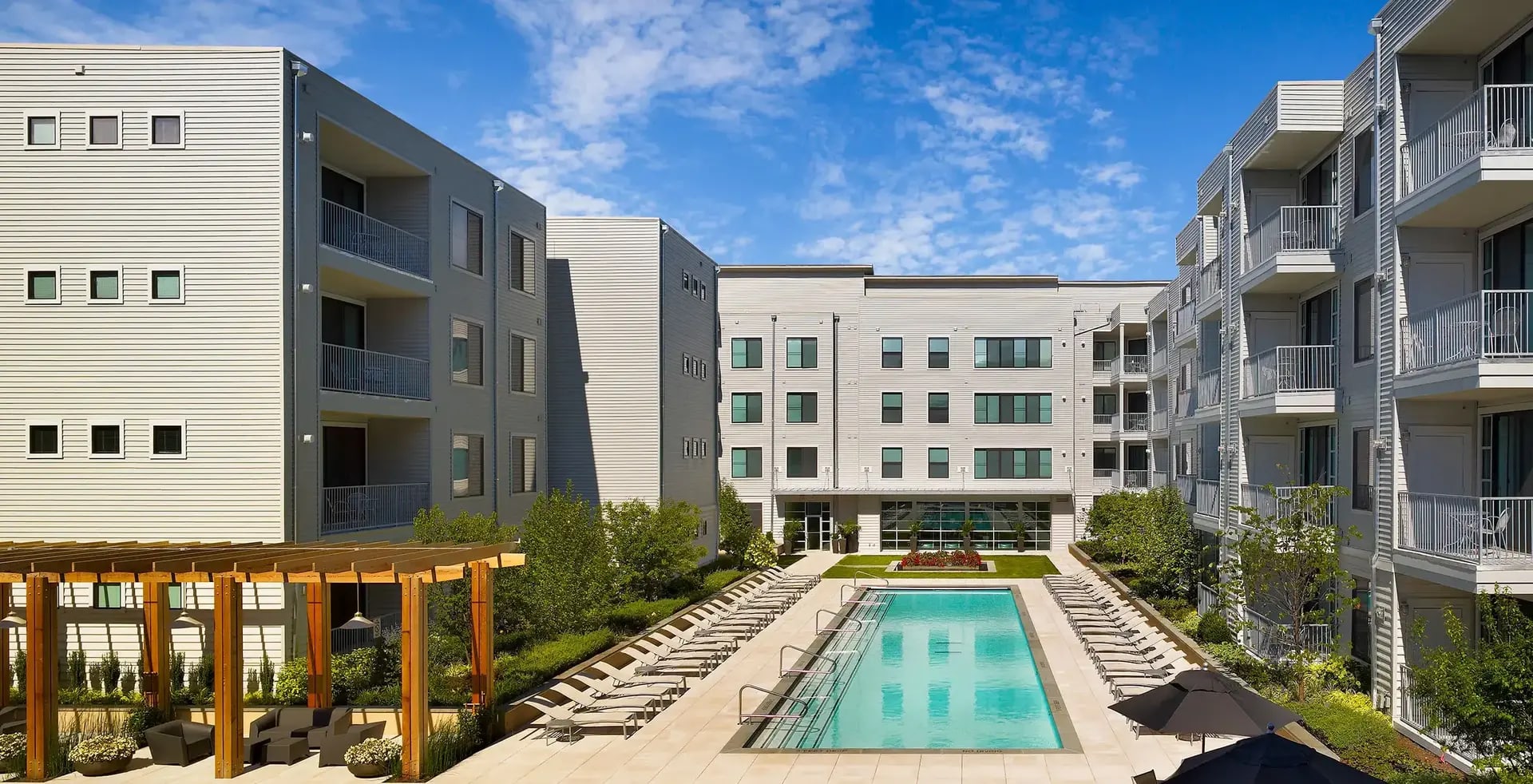 Courtyard view of multi-story apartment complex with rectangular pool, lounge chairs, wooden pergola, and surrounding residential buildings under blue sky.