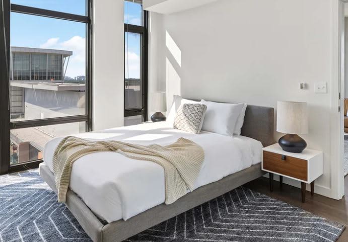 Bedroom with gray upholstered bed, white bedding, wood nightstand, and large windows overlooking Arlington cityscape.