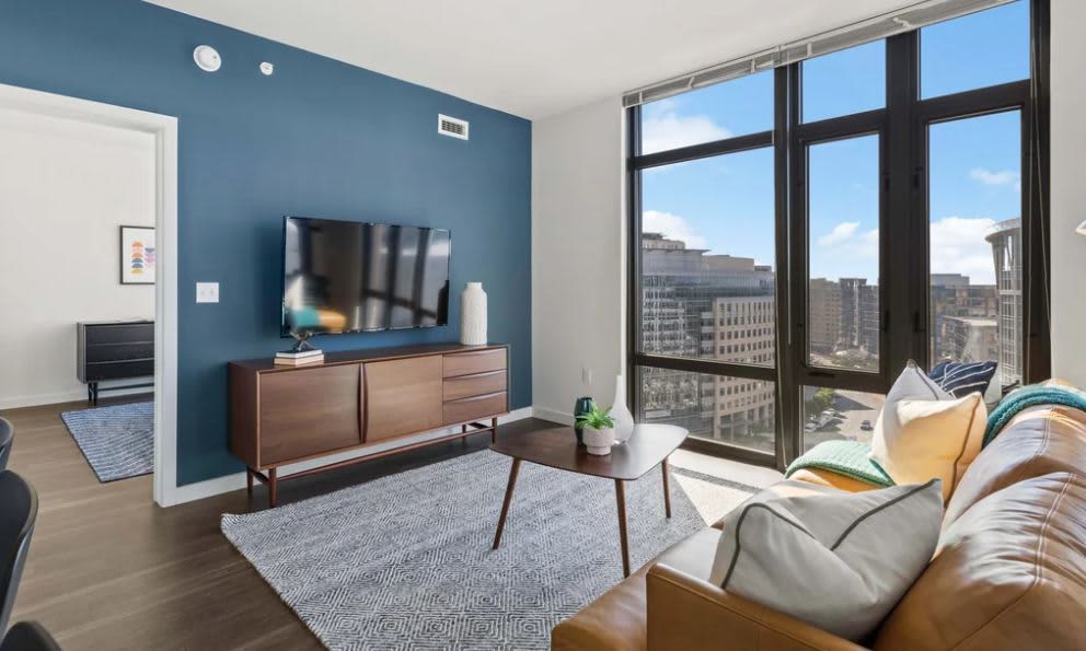 Living room with teal accent wall, wall-mounted TV on wooden media console, tan leather sofa, and expansive windows with downtown Arlington views.