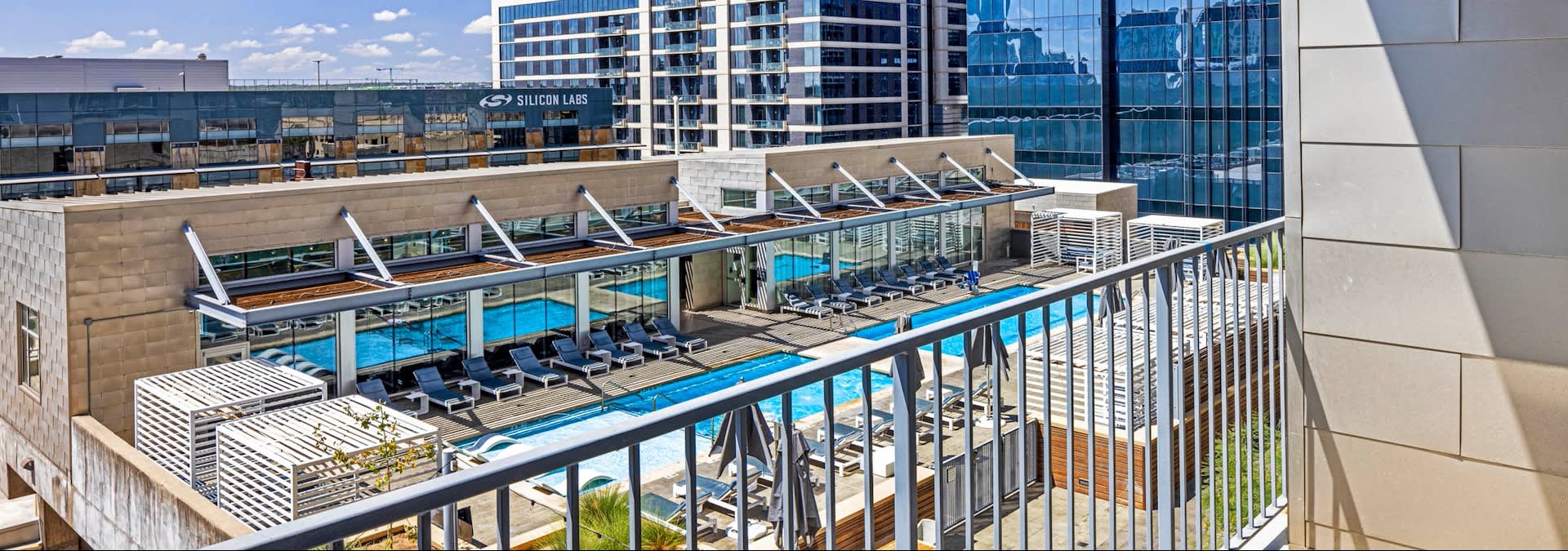 Rooftop pool deck with blue swimming pool, lounge chairs, umbrellas, and metal railing overlooking Austin skyline.