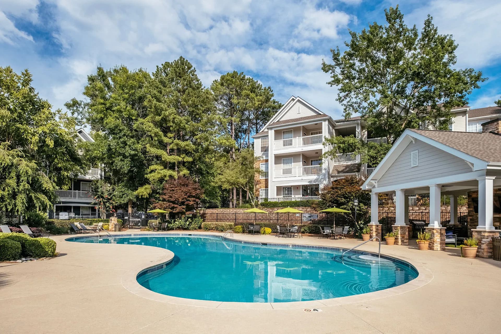 Resort-style swimming pool with curved shape and blue water, surrounded by concrete deck, white columned pavilion, and three-story residential buildings with white railings.