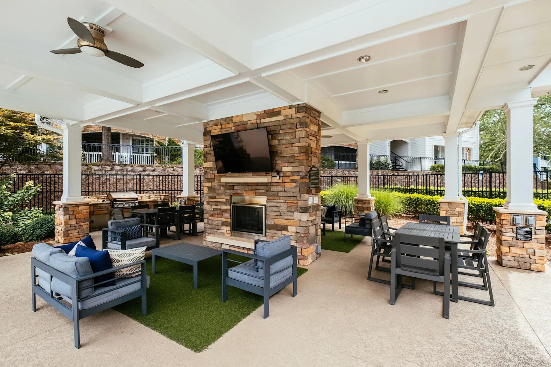 Covered patio with brick fireplace, mounted TV, gray outdoor furniture, dining table, and manicured landscaping beneath white coffered ceiling.