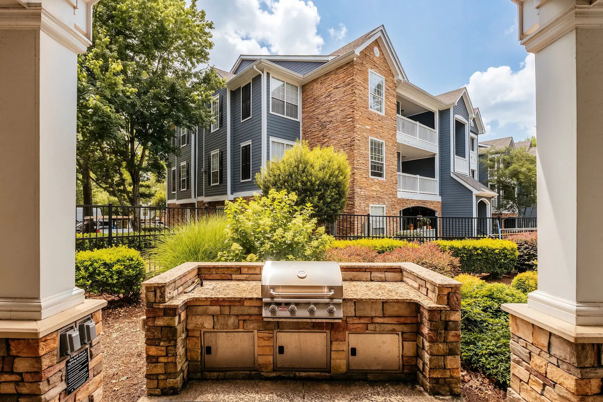 Outdoor kitchen with built-in gas grill and stone countertops, brick residential buildings visible in background with landscaped grounds.