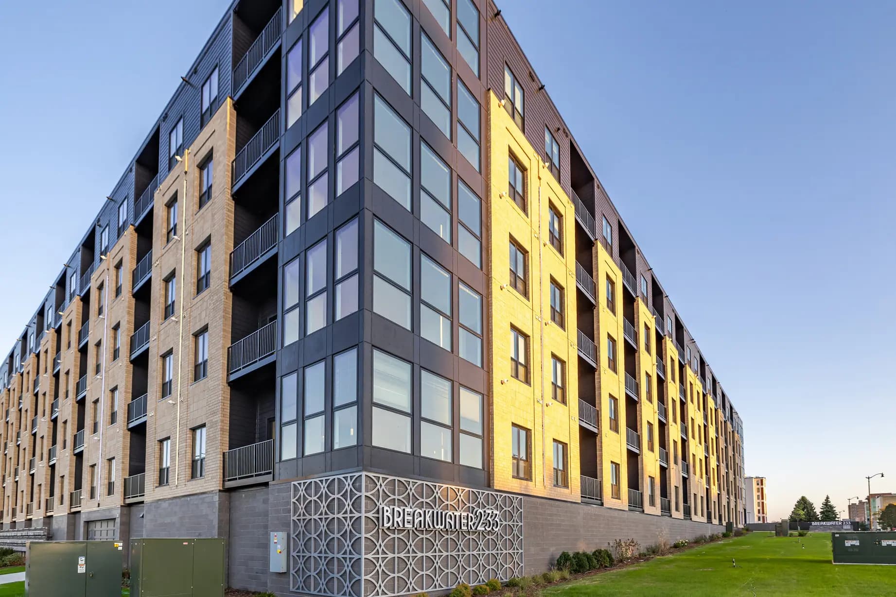 Multi-story residential building with tan brick, dark metal framing, and bright yellow facade sections, green lawn in foreground.