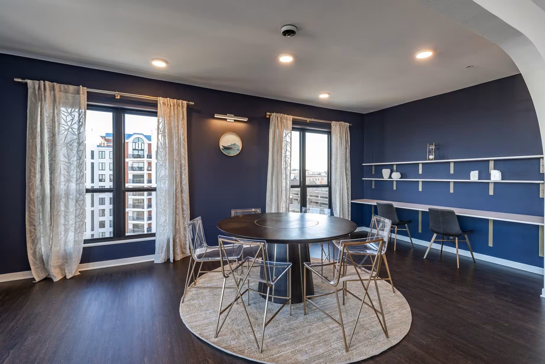 Dining room with round black table, four chairs, navy blue walls with floating shelves, large windows with beige curtains, and dark hardwood floors.