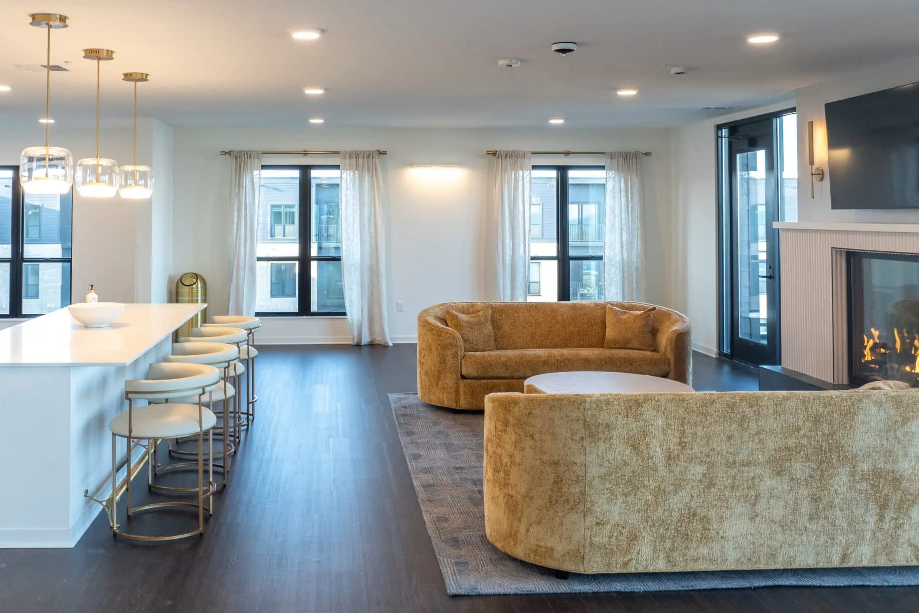 Open-concept living room with tan curved sofa, kitchen island with seating, dark wood floors, and gas fireplace with large windows overlooking the city.