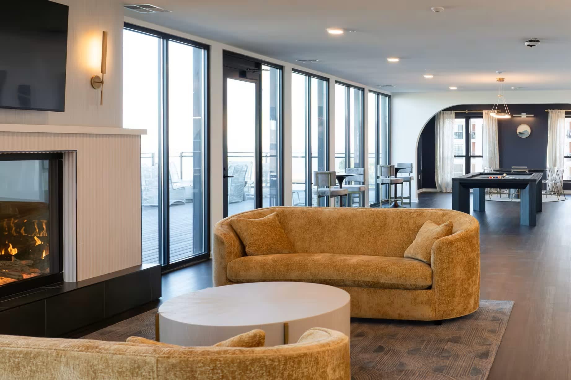 Modern living room with curved mustard sofa, white round coffee table, gas fireplace, and expansive glass doors opening to a waterfront balcony.