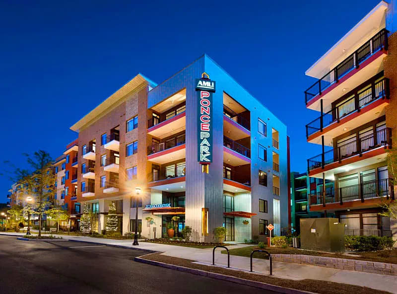 Multi-story apartment complex with orange, blue, and tan exterior, illuminated signage, ground-floor retail, and landscaped street at dusk.