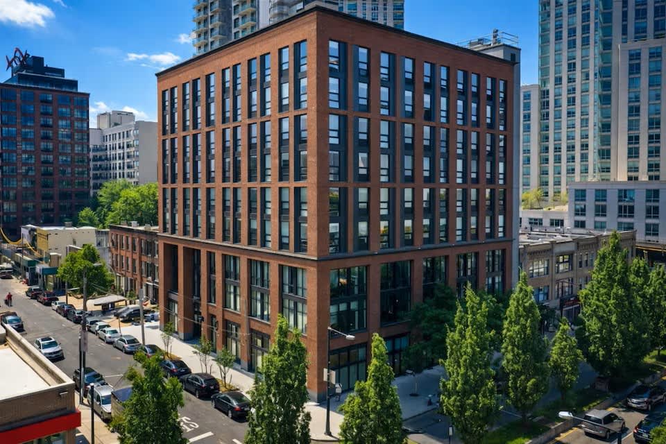 Brick residential building with regular window grid, ground-floor retail, green landscaping, and surrounding high-rise buildings under blue sky.