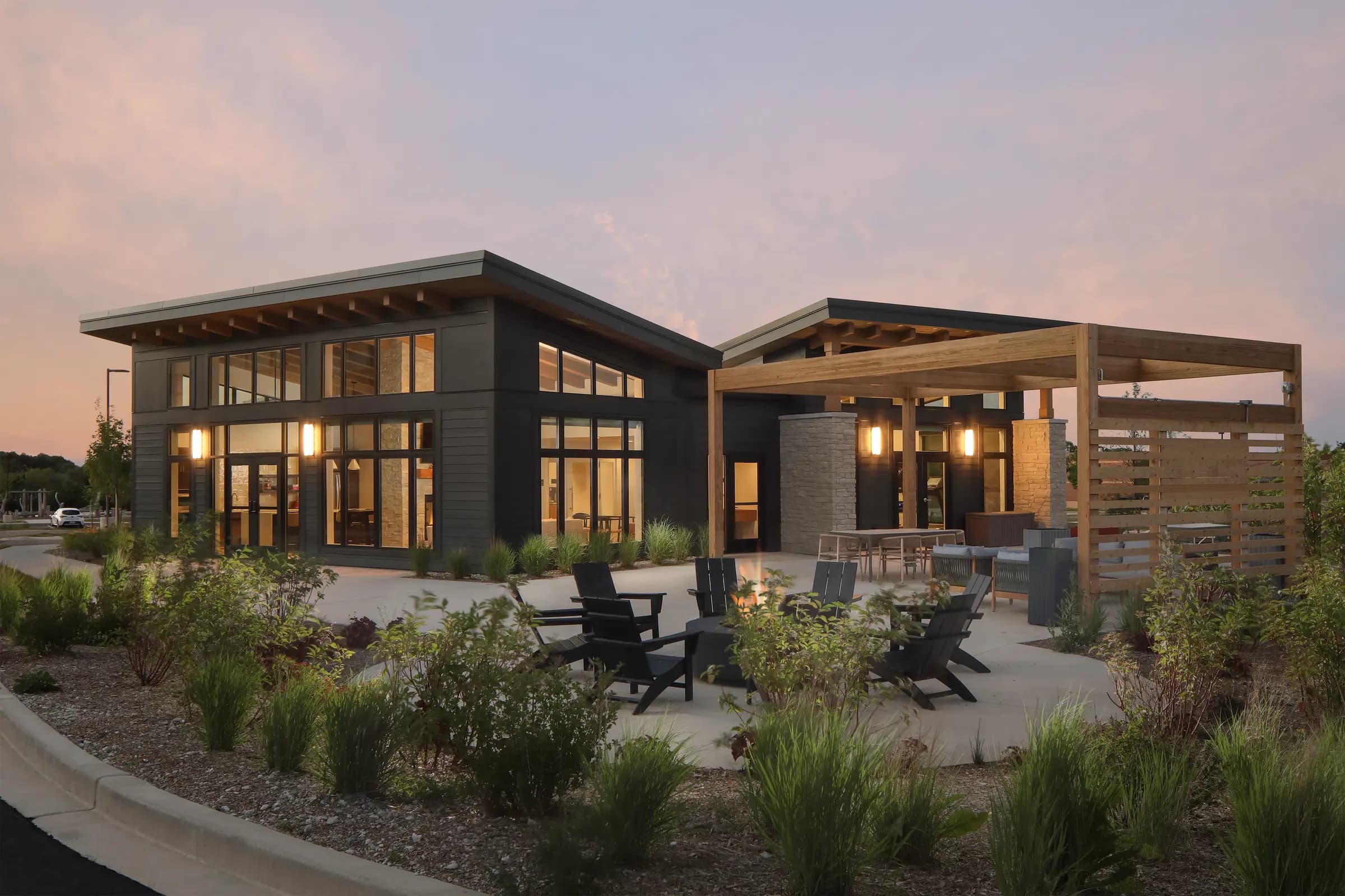 Modern residential building with dark exterior, extensive windows, wooden pergola, and patio seating at dusk.