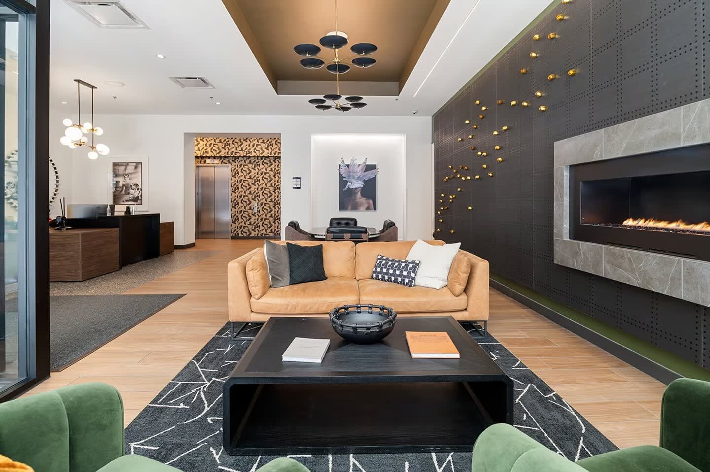Modern lounge with tan leather sofa, black fireplace wall with gold accent lights, wood flooring, and contemporary pendant lighting.