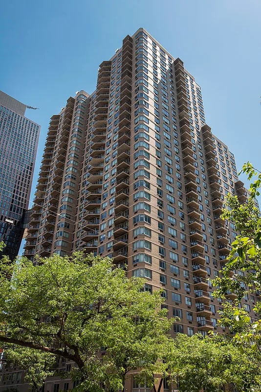 Tall residential high-rise building with brown brick facade, rows of balconies, and blue-tinted windows against clear sky, surrounded by green trees.