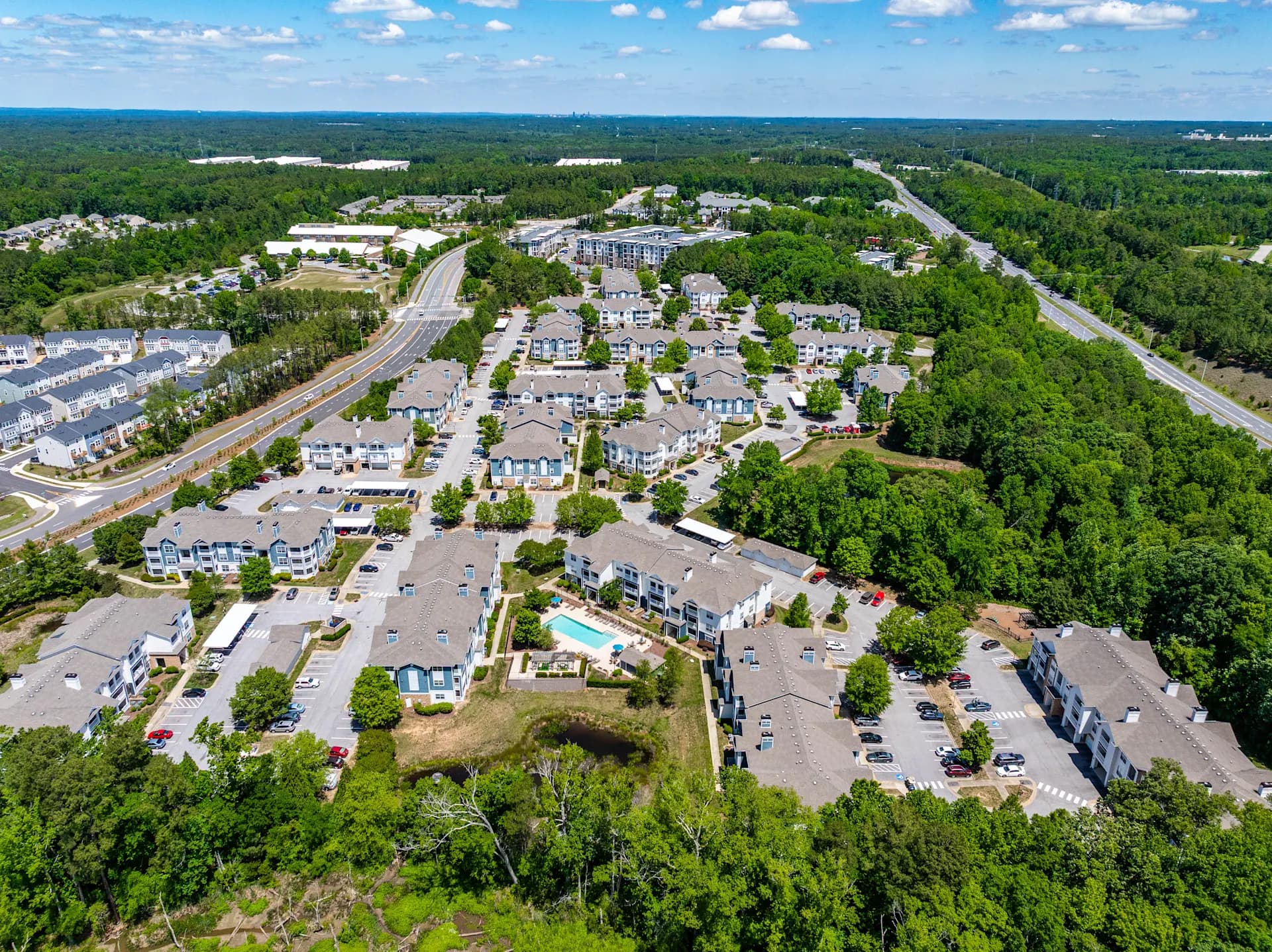 Aerial view of apartment complex nestled in green forest with parking lots, pool, and nearby highway in Cary, North Carolina.