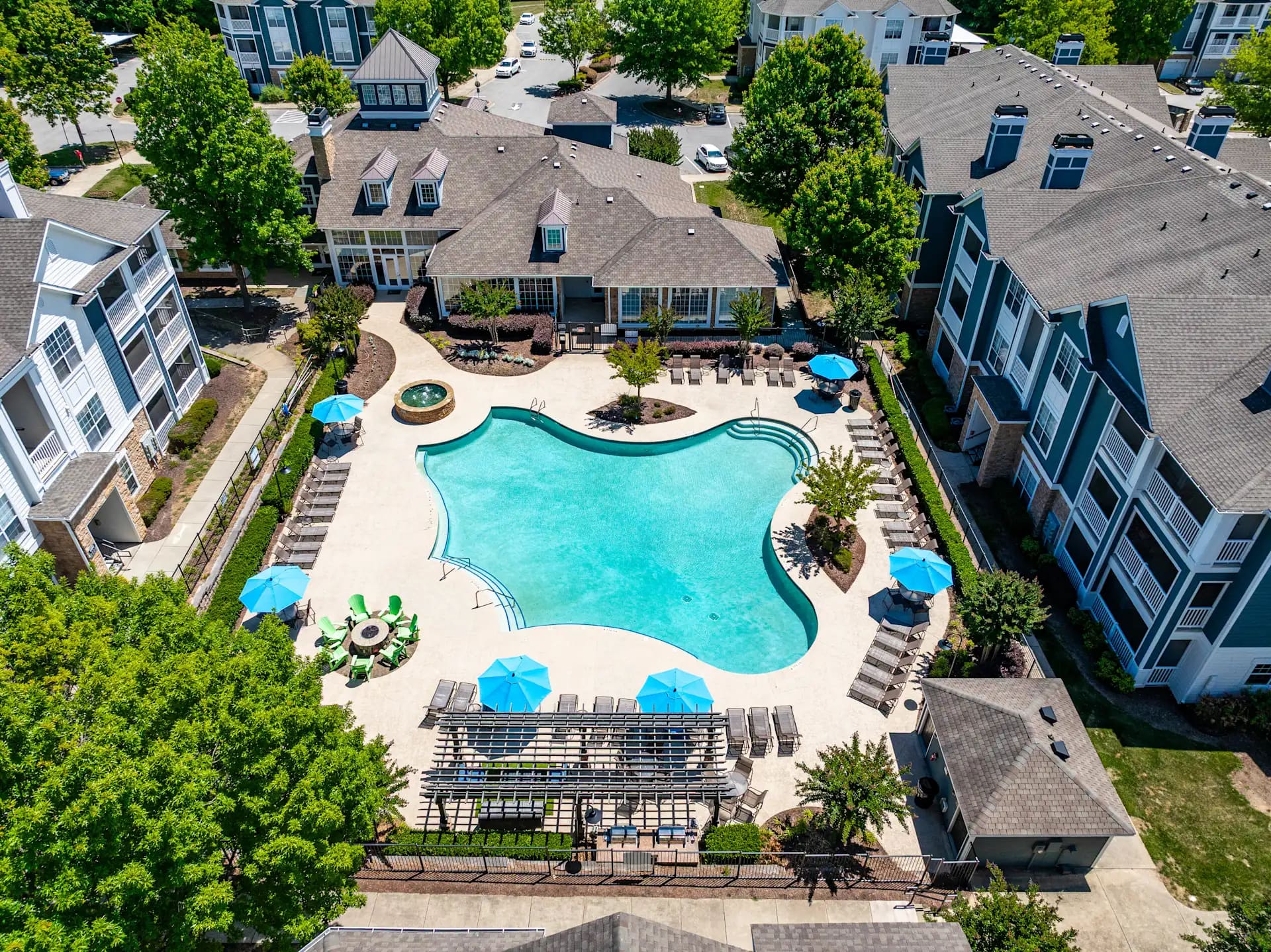 Aerial view of a curved swimming pool with blue umbrellas, lounge chairs, and landscaped courtyard surrounded by multi-story apartment buildings.