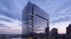 Modern glass high-rise building with vertical ribbed facade at dusk, surrounded by other city buildings under cloudy sky.