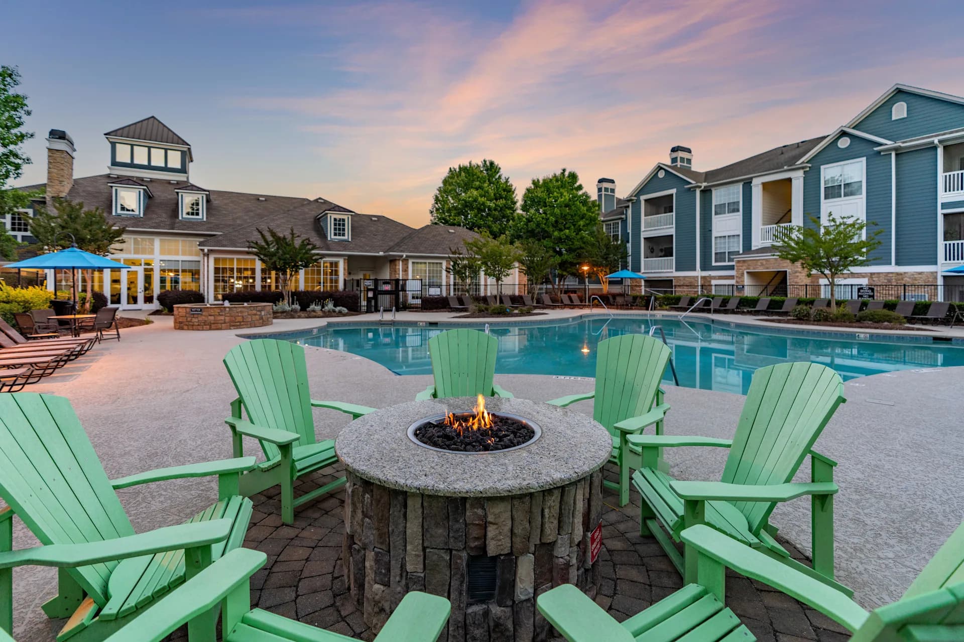 Resort-style pool area at dusk with green Adirondack chairs surrounding a lit fire pit, blue umbrellas, and multi-story apartment buildings in background.