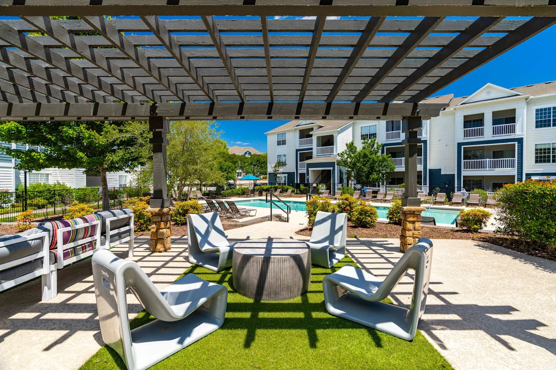 Outdoor lounge area under wooden pergola with modern white chairs, artificial grass, and swimming pool visible beyond landscaped grounds.