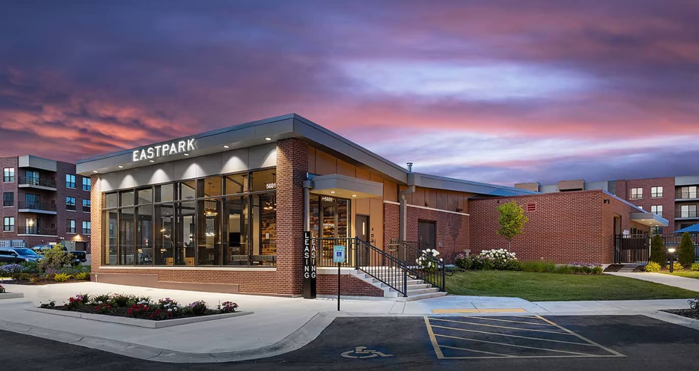 Modern brick leasing office building with glass storefront entrance, landscaping, and parking area at dusk.
