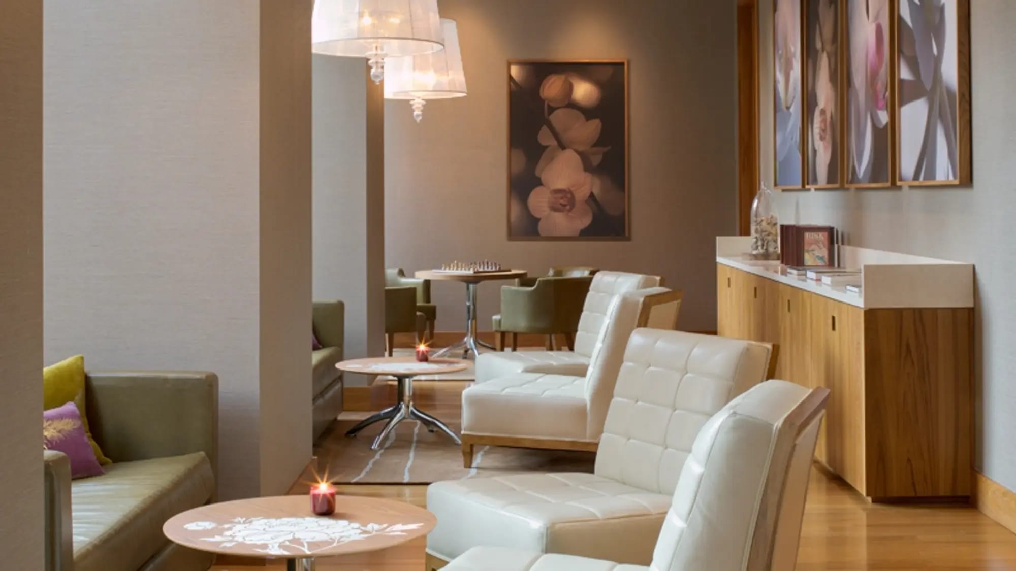 Modern lounge with cream quilted chairs, round side table, wood cabinetry, pendant lighting, and framed floral artwork on beige walls.