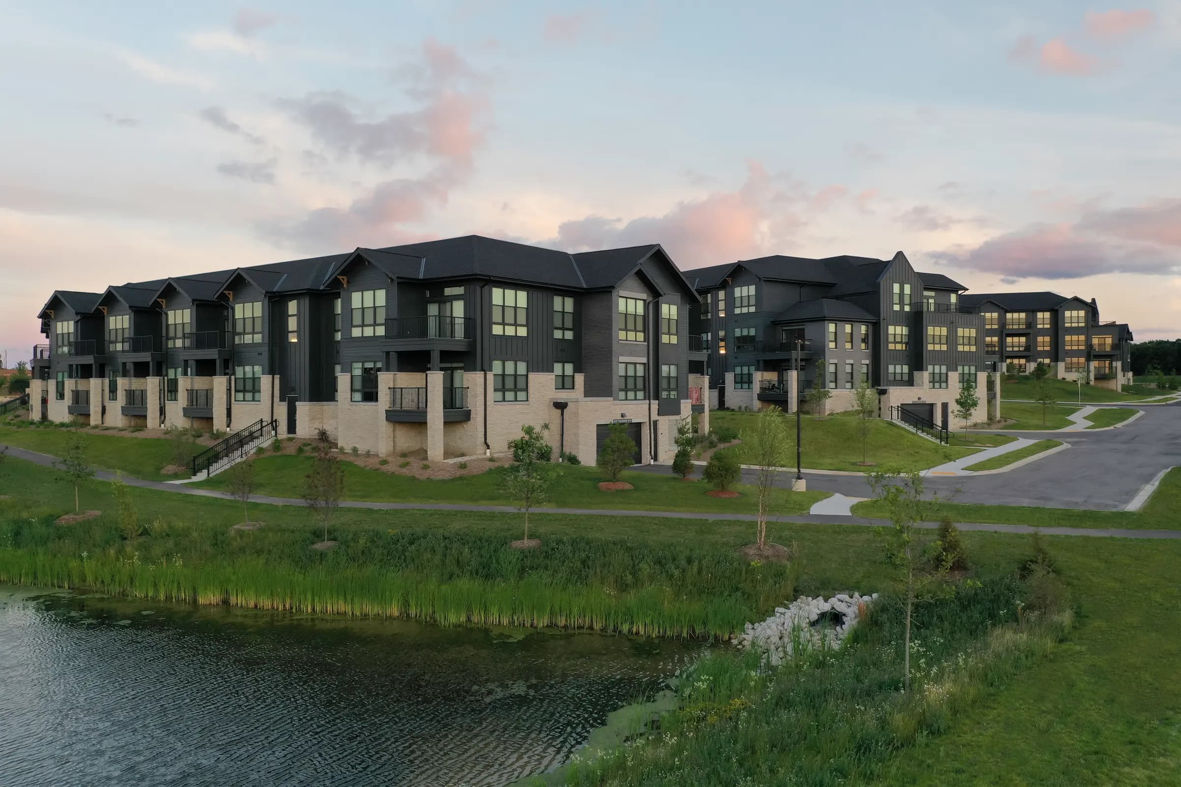 Multi-unit residential complex with dark siding and stone foundations facing a pond at dusk, landscaped grounds visible.