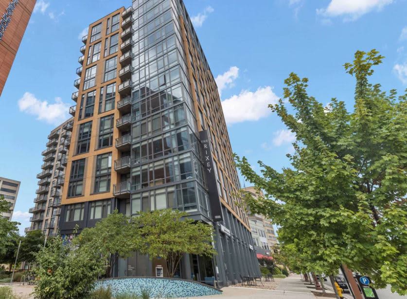 Tall mixed-use building with brick and glass facade, ground-level retail, and mature trees in Arlington streetscape.