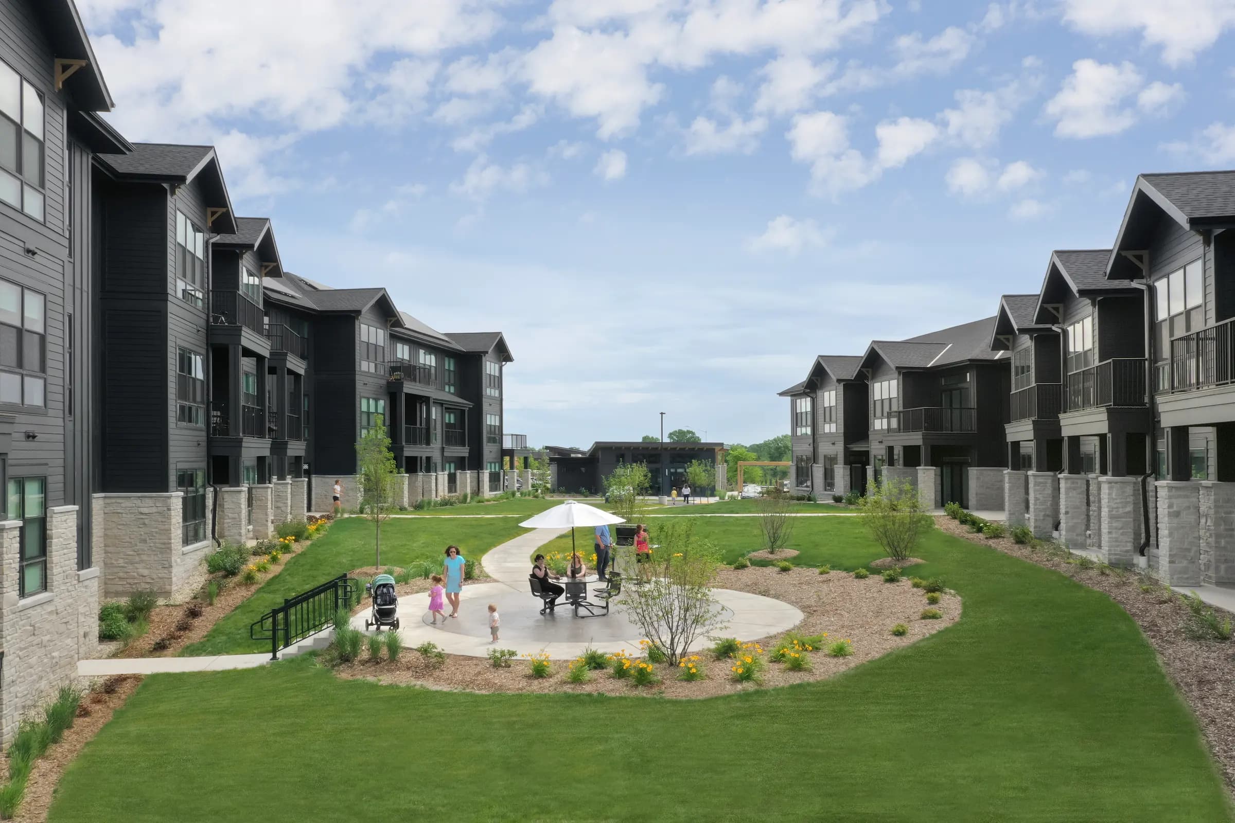 Central courtyard at Broadacre Apartments with manicured lawn, paved pathways, shade umbrella, and families enjoying the outdoor common space between multi-story residential buildings.