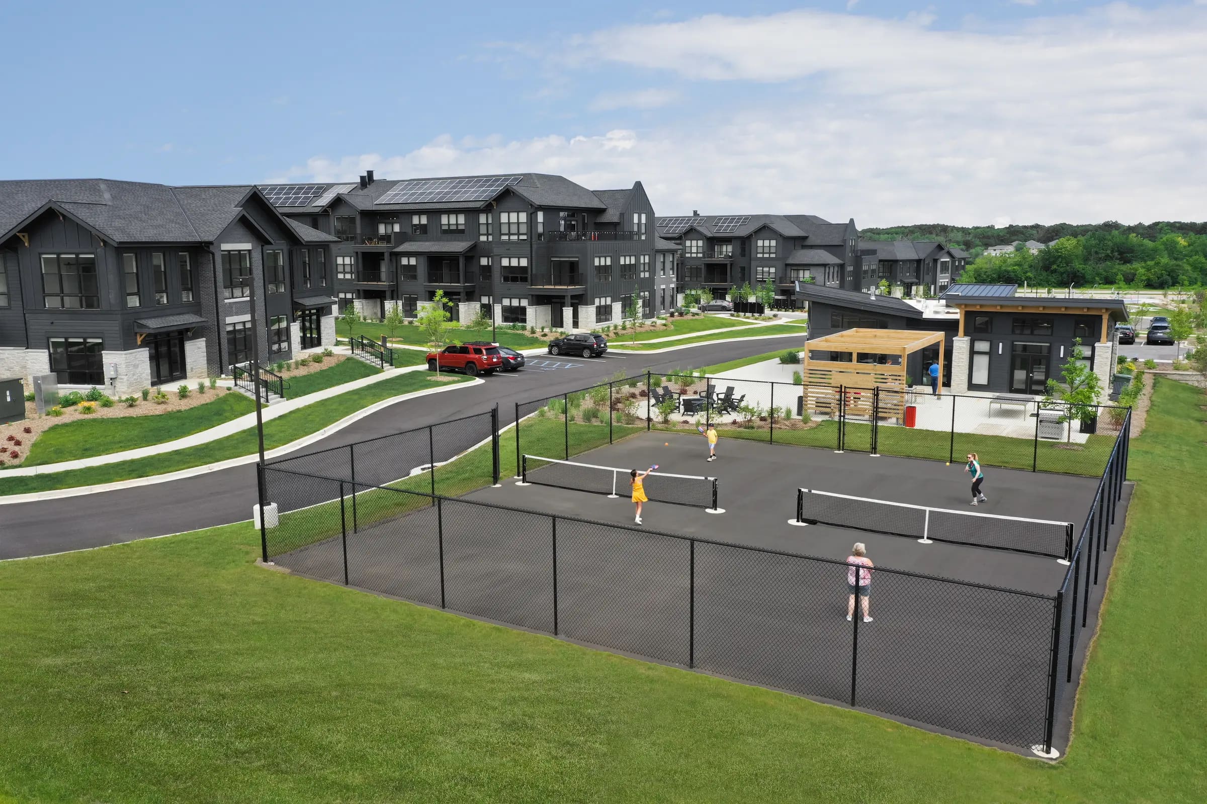 Fenced outdoor sports court with multiple courts, people playing, surrounded by modern dark-exterior residential buildings and green lawn.
