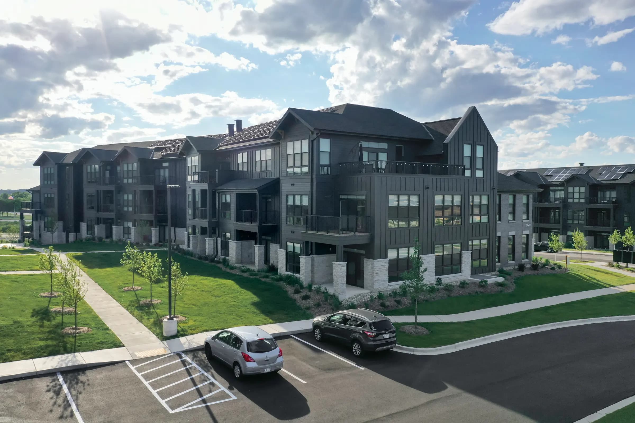 Multi-unit residential building with dark siding and white brick lower level, parked cars in foreground, green lawn and young trees visible.