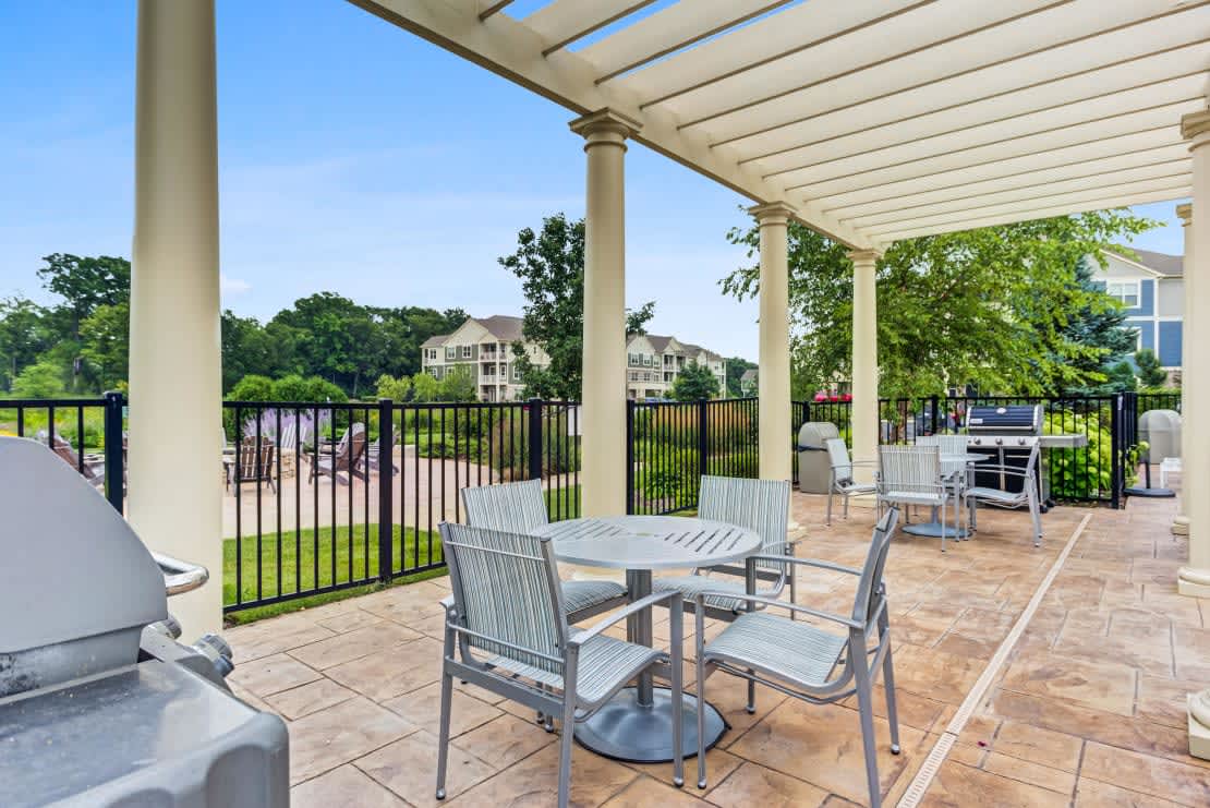 Rooftop terrace with white pergola, metal dining chairs, round table, black railing, and views of residential buildings and green landscape.