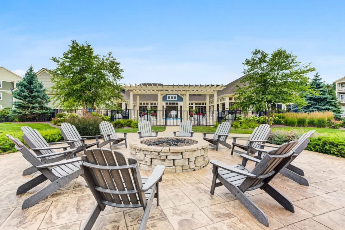 Outdoor courtyard with multiple gray Adirondack chairs arranged around a circular stone fire pit, with a pergola-topped clubhouse and green lawn in the background.