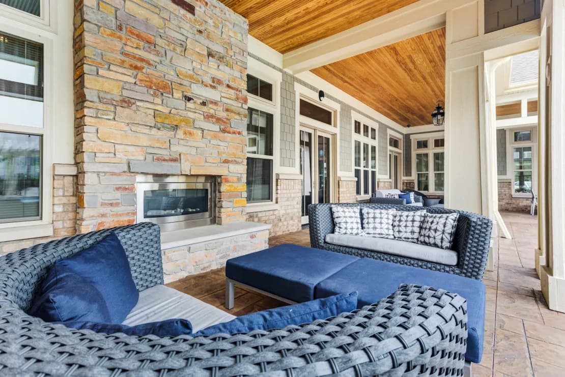 Covered patio with woven lounge chairs, blue cushions, brick fireplace, and wood ceiling overlooking additional seating areas.